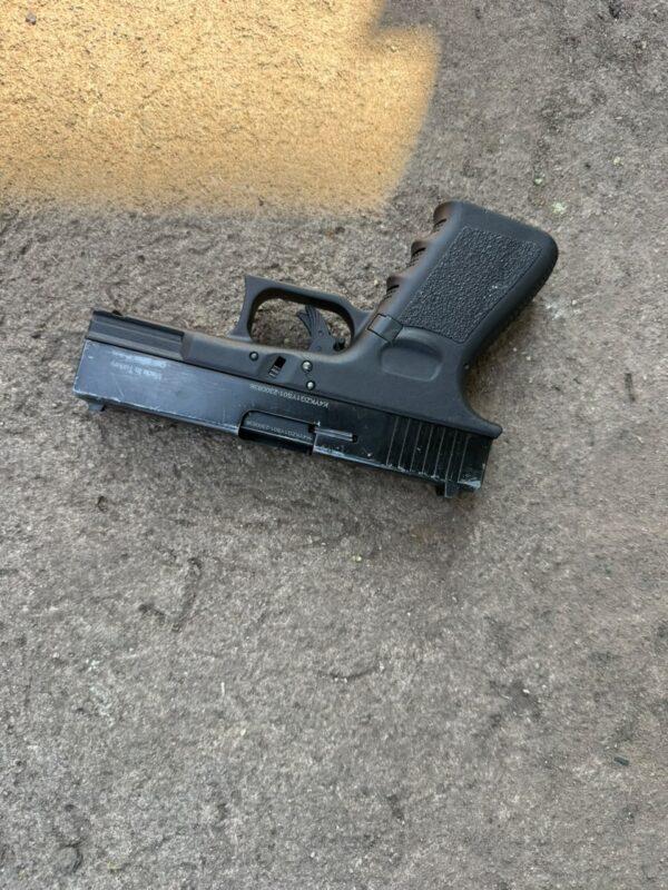 Black semi-automatic handgun lying on rough concrete pavement, viewed from above, with sunlight and shadows creating a stark, somber scene.
