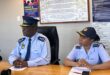 Two police officers sit at a wooden table beneath an anti-corruption pledge and code of conduct posters, conveying professionalism and integrity.
