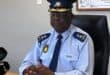 Police officer in uniform and peaked cap sits at a desk during a serious press briefing, with microphones and an “OUT! Fraud & Corruption” poster.
