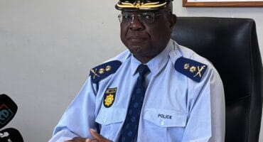 Police officer in uniform and peaked cap sits at a desk during a serious press briefing, with microphones and an “OUT! Fraud & Corruption” poster.