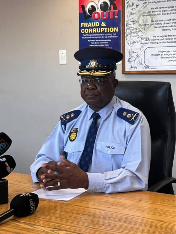 Police officer in formal uniform sits at a desk before microphones, with a serious expression; a poster warns of fraud and corruption.