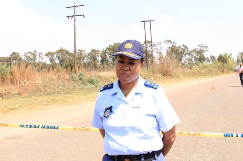 Police officer in uniform stands behind caution tape on a rural road, with utility poles, dry grass, and trees in the background.
