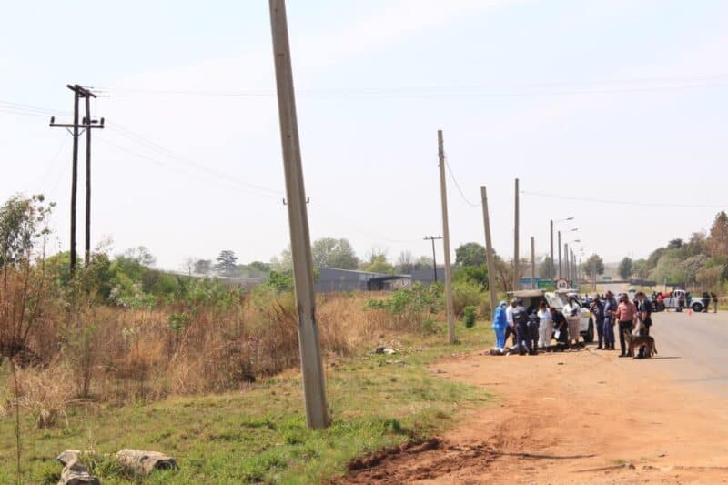 A group of people gathers beside a dusty rural road lined with utility poles, dry vegetation, and scattered homes under a hazy sky.