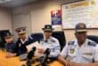 Four uniformed police officials sit at a conference table with microphones; one reads a document, with an anti-corruption pledge poster behind them.