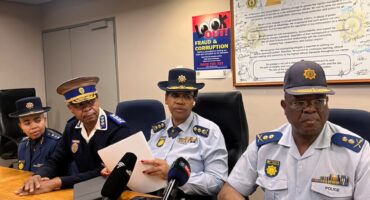 Four uniformed police officials sit at a conference table with microphones; one reads a document, with an anti-corruption pledge poster behind them.