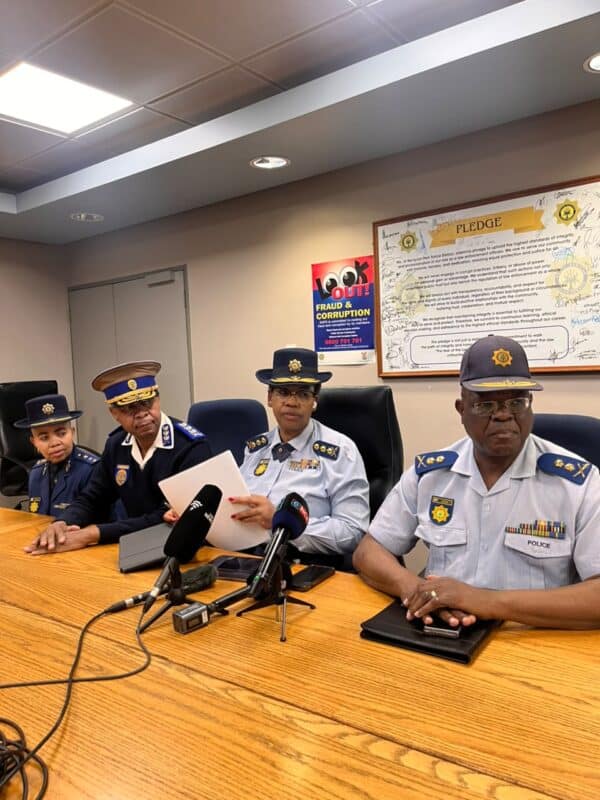 Four uniformed police officials sit at a conference table with microphones; one reads a document, with an anti-corruption pledge poster behind them.