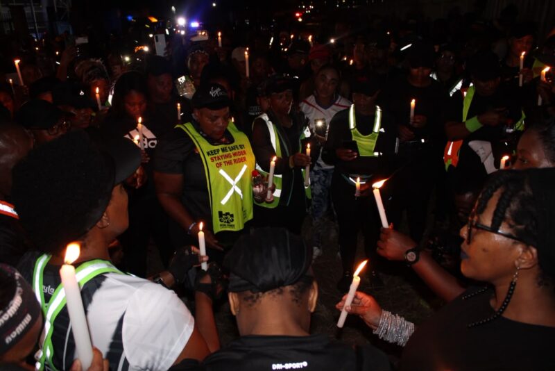 Crowd gathers at a nighttime candlelight vigil, holding candles; volunteers in reflective vests stand among mourners in a solemn scene.