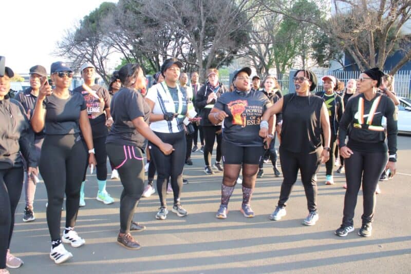 Diverse women in athletic wear gather for a community walk on a tree-lined street, smiling and chatting in bright evening light.