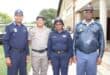 Four uniformed police officers pose outside a building, smiling at the camera; two women and two men wear navy, gray, and blue uniforms.