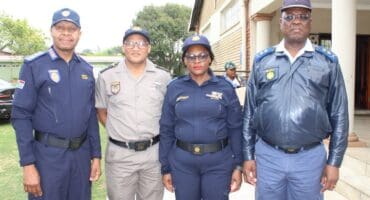 Four uniformed police officers pose outside a building, smiling at the camera; two women and two men wear navy, gray, and blue uniforms.