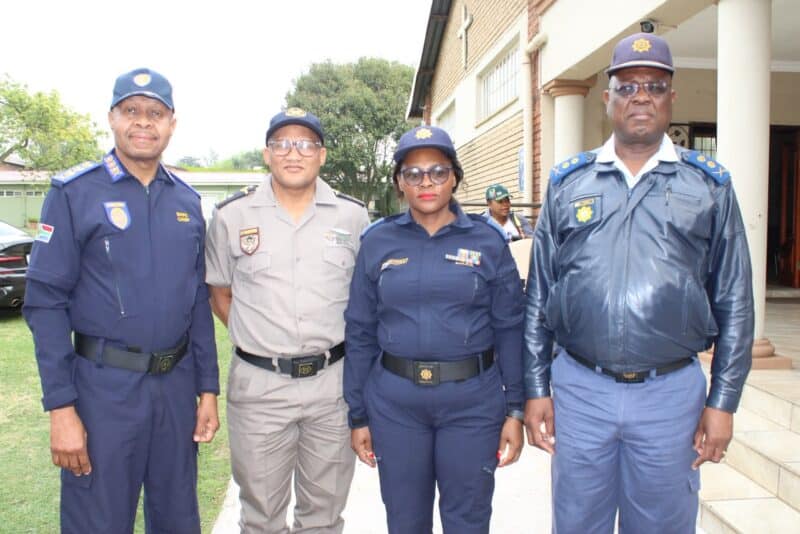Four uniformed police officers pose outside a building, smiling at the camera; two women and two men wear navy, gray, and blue uniforms.