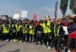 A diverse crowd in high-visibility vests rallies on a city street, holding signs and flags, with linked arms conveying solidarity and determination.