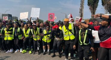A diverse crowd in high-visibility vests rallies on a city street, holding signs and flags, with linked arms conveying solidarity and determination.