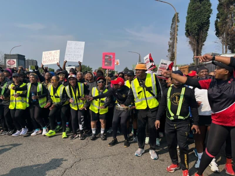 A diverse crowd in high-visibility vests rallies on a city street, holding signs and flags, with linked arms conveying solidarity and determination.