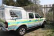 South African Police Service vehicles—a marked police pickup and van—parked beside a fence in daylight, conveying an official, utilitarian mood.