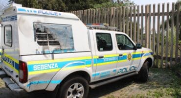 South African Police Service vehicles—a marked police pickup and van—parked beside a fence in daylight, conveying an official, utilitarian mood.