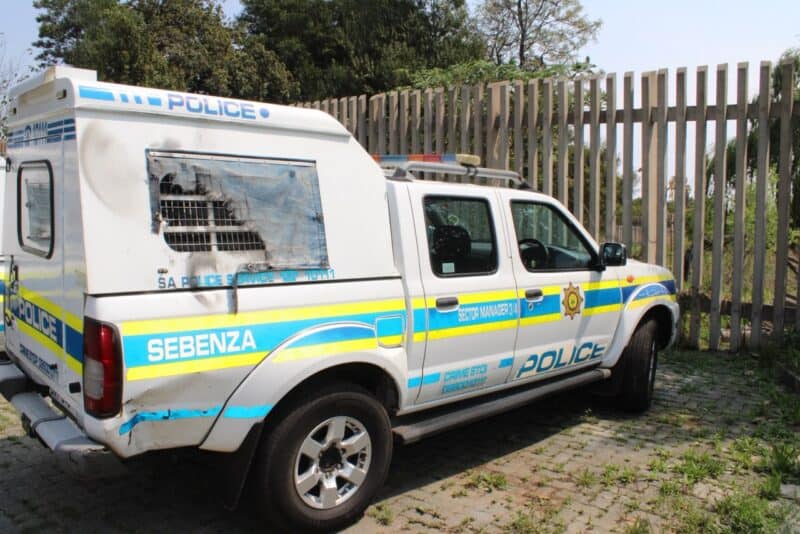 South African Police Service vehicles—a marked police pickup and van—parked beside a fence in daylight, conveying an official, utilitarian mood.