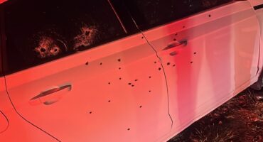 At night, red and blue emergency lights illuminate a white sedan peppered with apparent bullet holes as an officer examines it roadside.