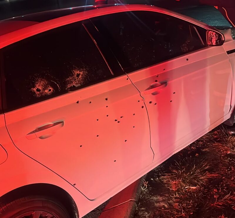 At night, red and blue emergency lights illuminate a white sedan peppered with apparent bullet holes as an officer examines it roadside.
