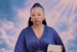 Portrait of a poised Black woman in a pleated blue dress holding a beige clutch, framed by dreamy pink clouds and radiant sunlight.