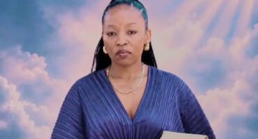 Portrait of a poised Black woman in a pleated blue dress holding a beige clutch, framed by dreamy pink clouds and radiant sunlight.