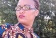 Portrait of a woman with braided hair and glasses wearing a colorful patterned outfit, posed outdoors against leafy trees with a calm expression.
