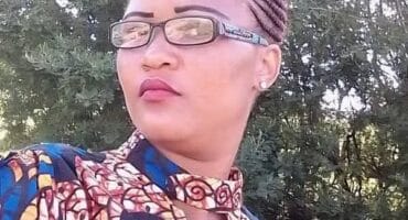 Portrait of a woman with braided hair and glasses wearing a colorful patterned outfit, posed outdoors against leafy trees with a calm expression.