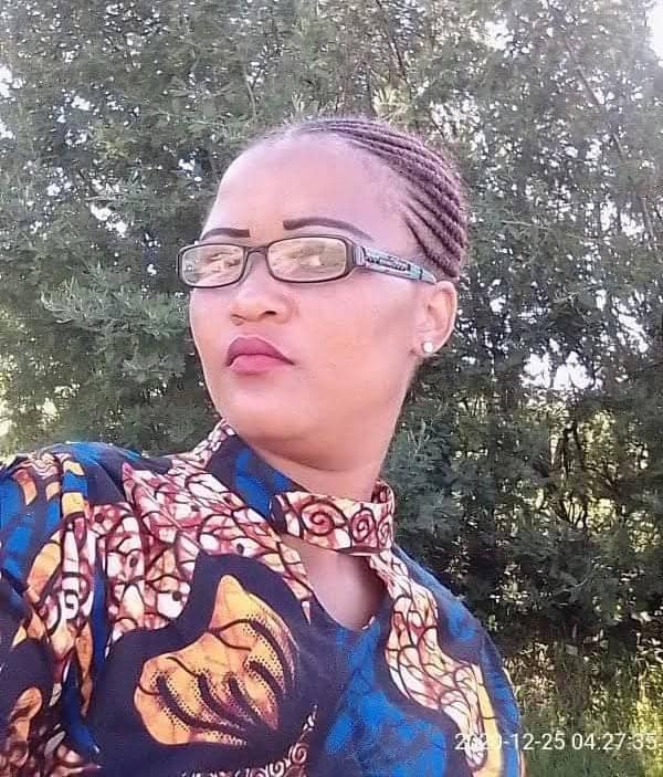 Portrait of a woman with braided hair and glasses wearing a colorful patterned outfit, posed outdoors against leafy trees with a calm expression.