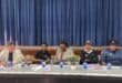 Five women sit on a panel behind a table with water bottles and flowers, including two in dark uniforms, against blue stage curtains.
