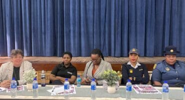Five women sit on a panel behind a table with water bottles and flowers, including two in dark uniforms, against blue stage curtains.