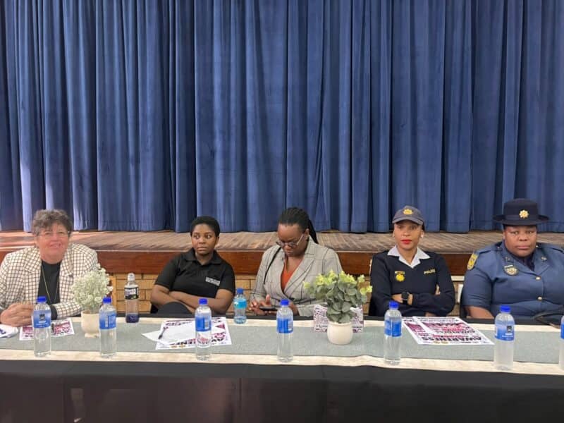 Five women sit on a panel behind a table with water bottles and flowers, including two in dark uniforms, against blue stage curtains.