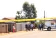 Yellow police tape blocks a sunny residential street as officers and bystanders gather beside a police van, creating a tense crime-scene atmosphere.