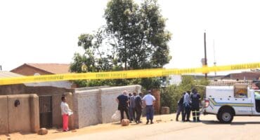 Yellow police tape blocks a sunny residential street as officers and bystanders gather beside a police van, creating a tense crime-scene atmosphere.