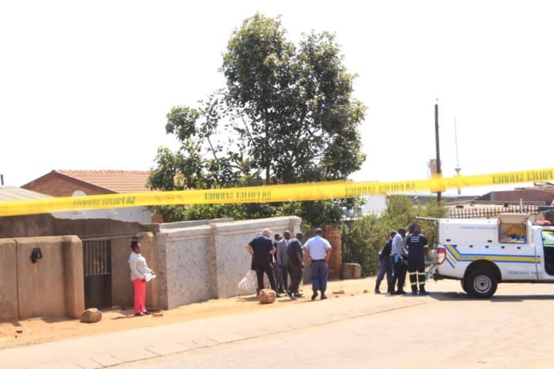 Yellow police tape blocks a sunny residential street as officers and bystanders gather beside a police van, creating a tense crime-scene atmosphere.