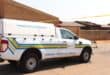 A white forensic pathology service pickup with blue and yellow stripes is parked beside a brick building in bright sunlight, conveying a somber, official tone.