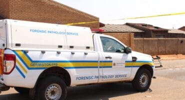 A white forensic pathology service pickup with blue and yellow stripes is parked beside a brick building in bright sunlight, conveying a somber, official tone.