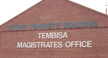 A red-brick gabled facade displays blue “Steve Tshwete Building” and white “Tembisa Magistrates Office” lettering, conveying a formal civic setting.
