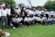 A large group of adults in white golf shirts and caps pose on a grassy field beneath event flags, smiling in a cheerful, team-oriented gathering.