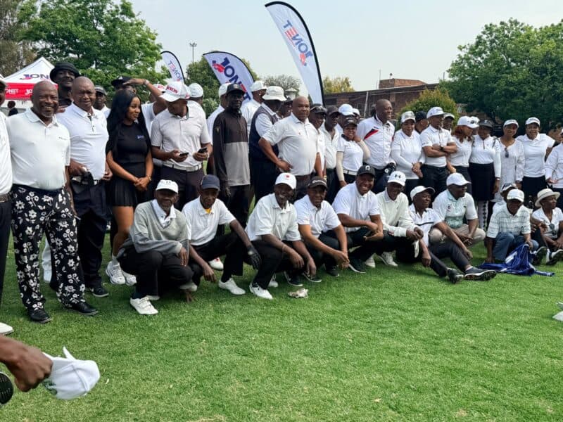 A large group of adults in white golf shirts and caps pose on a grassy field beneath event flags, smiling in a cheerful, team-oriented gathering.