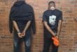 Two men stand facing a brick wall with hands cuffed behind their backs; a no-smoking sign hangs above, creating a somber, restrained scene.
