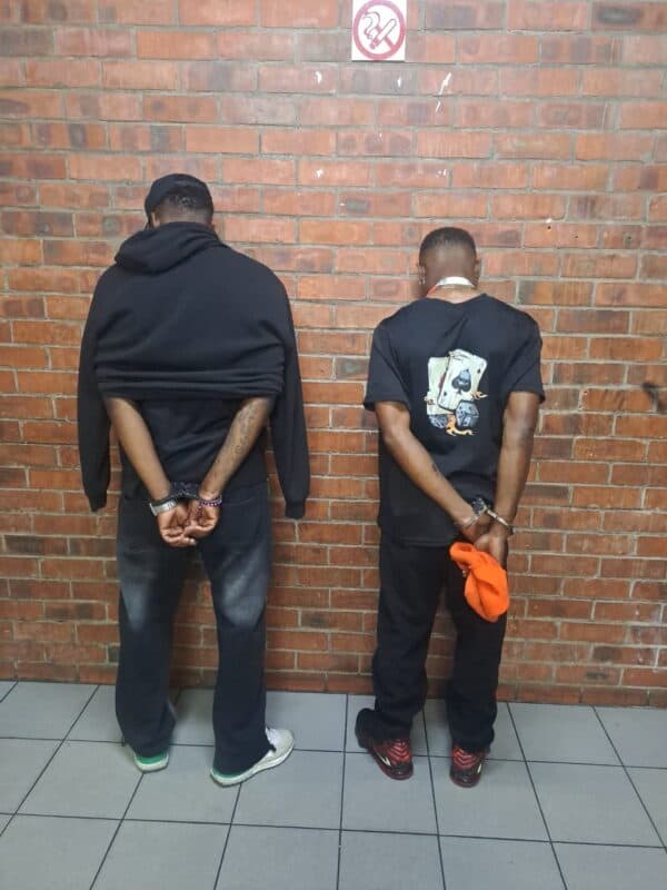 Two men stand facing a brick wall with hands cuffed behind their backs; a no-smoking sign hangs above, creating a somber, restrained scene.