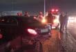 At night, a black sedan with a crushed rear bumper sits on gravel as responders stand near flashing emergency vehicles, creating a tense scene.