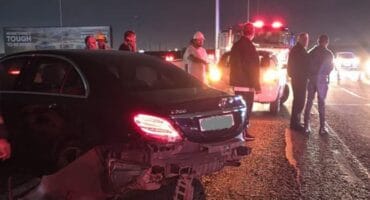 At night, a black sedan with a crushed rear bumper sits on gravel as responders stand near flashing emergency vehicles, creating a tense scene.