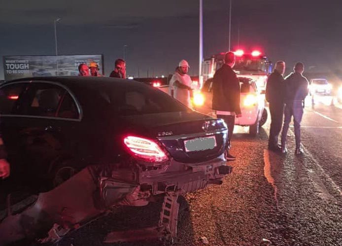At night, a black sedan with a crushed rear bumper sits on gravel as responders stand near flashing emergency vehicles, creating a tense scene.
