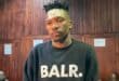 A young man with a bandage on his forehead stands before a wood-paneled wall, wearing a black “BALR.” sweatshirt; his expression is subdued.