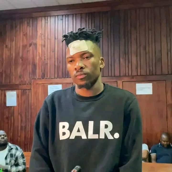 A young man with a bandage on his forehead stands before a wood-paneled wall, wearing a black “BALR.” sweatshirt; his expression is subdued.