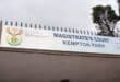 Sign above a gated entrance reads “Magistrate’s Court, Kempton Park,” beside South African government emblems; a tree rises against an overcast sky.