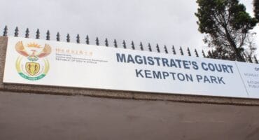 Sign above a gated entrance reads “Magistrate’s Court, Kempton Park,” beside South African government emblems; a tree rises against an overcast sky.