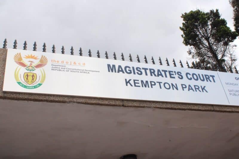 Sign above a gated entrance reads “Magistrate’s Court, Kempton Park,” beside South African government emblems; a tree rises against an overcast sky.