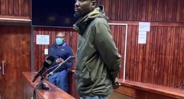 A serious-looking man in an olive jacket stands with hands behind his back in a wood-paneled courtroom, near microphones and a masked official.
